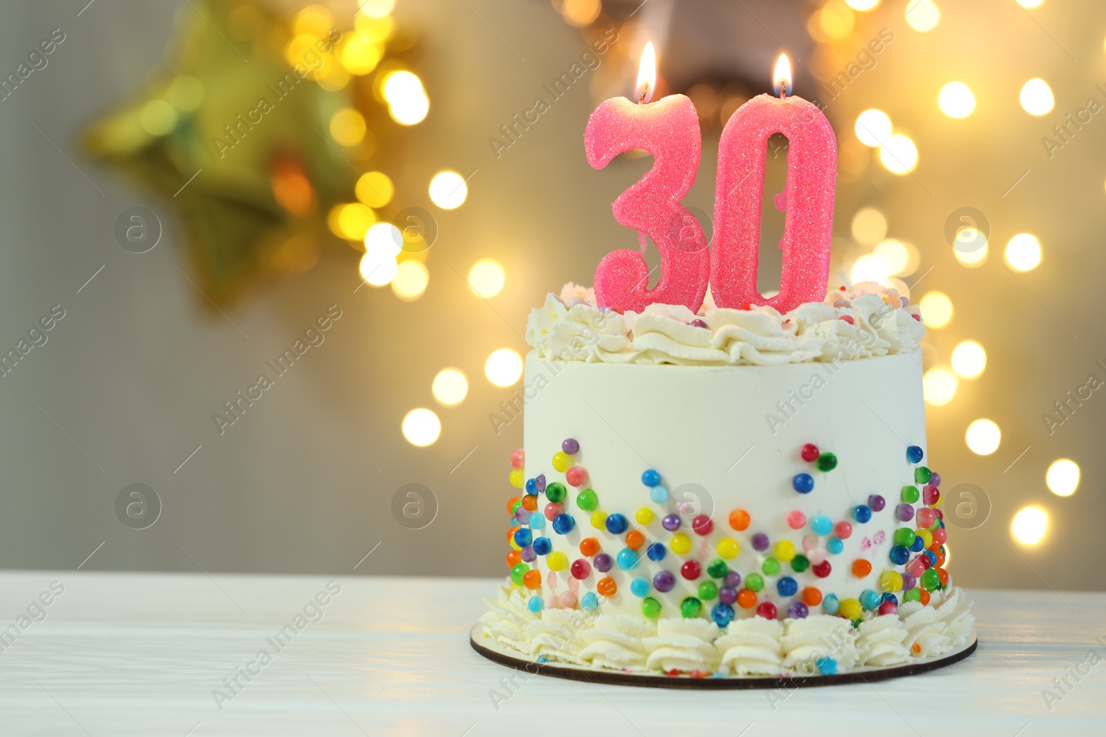 Photo of 30th birthday. Cake with burning number shaped candles on white wooden table against blurred background with lights, closeup and space for text. Bokeh effect