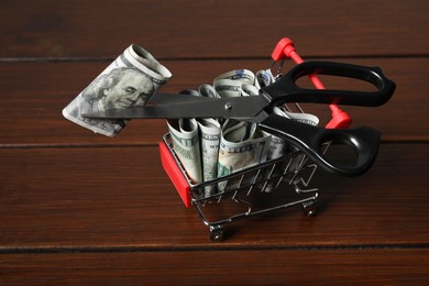 Photo of Dollar banknotes, scissors and small shopping cart on wooden table