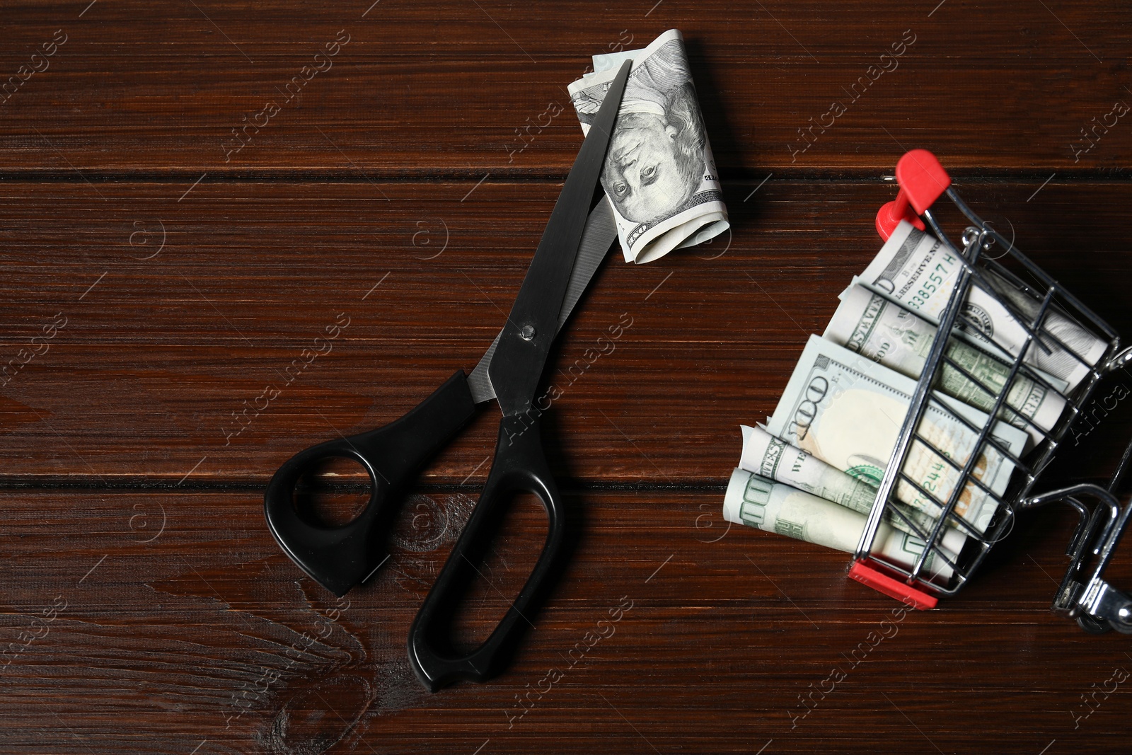 Photo of Dollar banknotes, scissors and small shopping cart on wooden table, flat lay