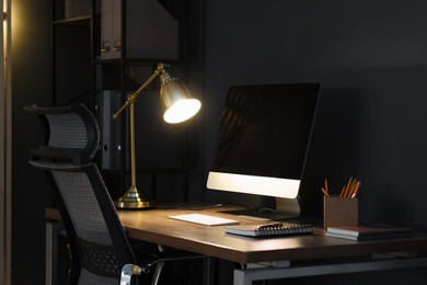 Photo of Comfortable workplace with computer and lamp on wooden desk at home office in evening