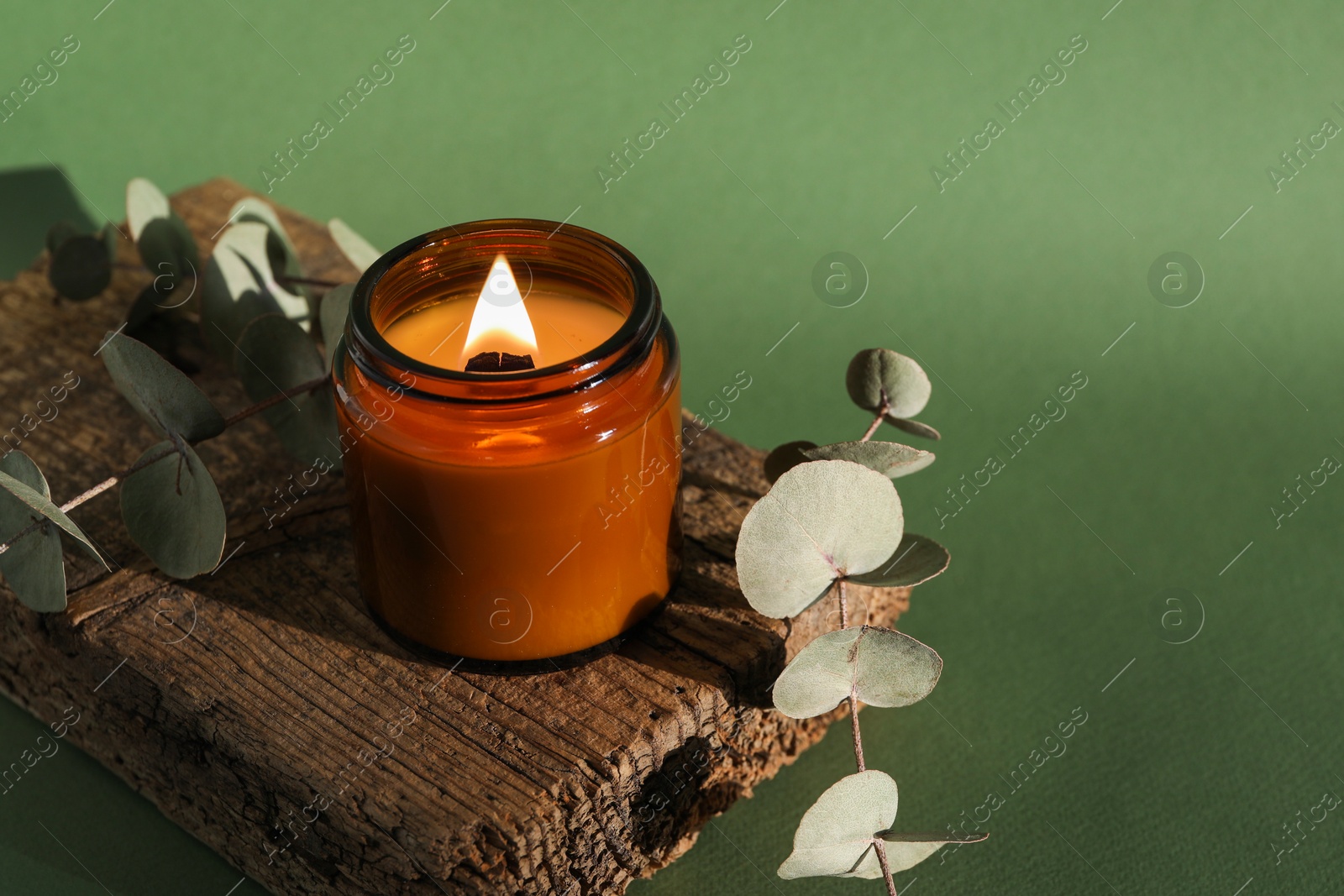 Photo of Burning soy wax candle, decorative piece of wood and eucalyptus branches on green background, space for text