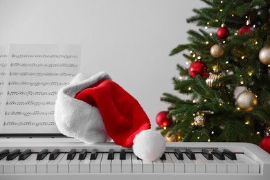 Photo of Synthesizer with music sheets and Santa hat near Christmas tree indoors, closeup