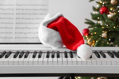 Photo of Synthesizer with music sheets and Santa hat near Christmas tree indoors, closeup