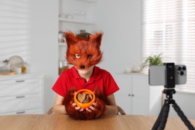 Photo of Quadrobics. Boy wearing fox mask and gloves with feeding bowl filming video at table indoors