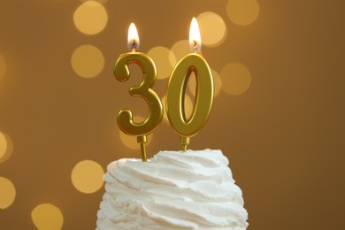 Photo of 30th birthday. Burning number shaped candles on cupcake against golden background with blurred lights, closeup. Bokeh effect