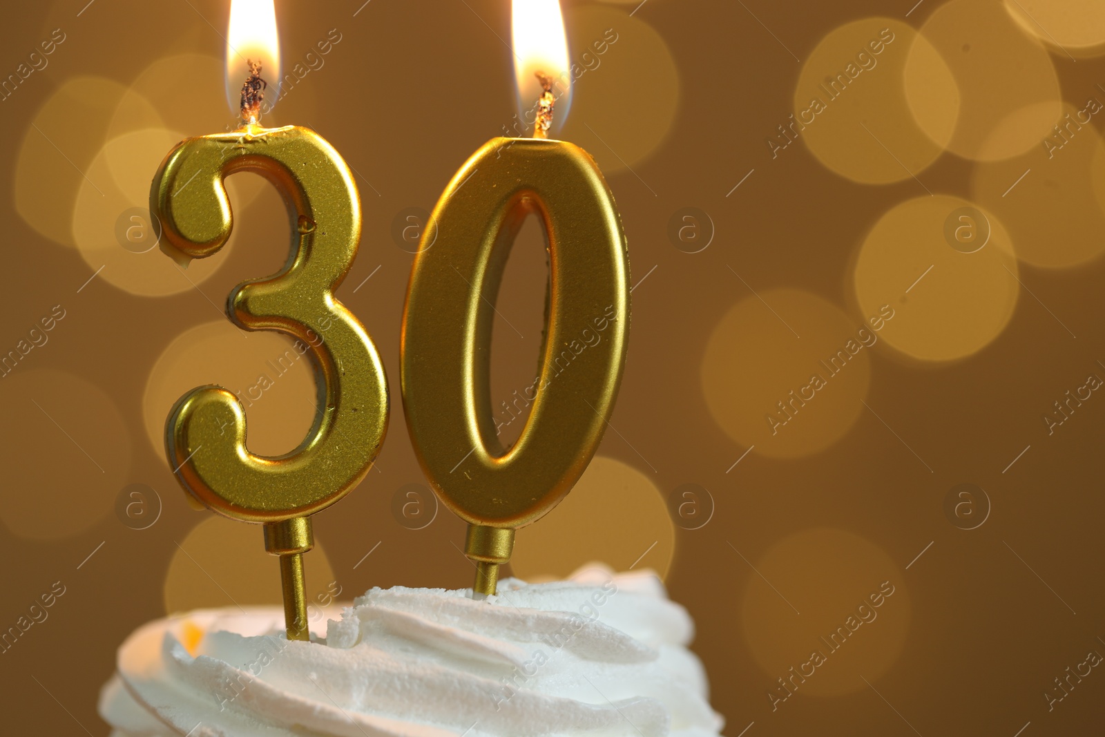 Photo of 30th birthday. Burning number shaped candles on cupcake against golden background with blurred lights, closeup and space for text. Bokeh effect