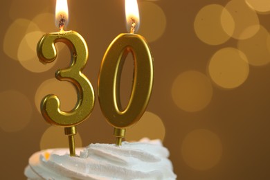 Photo of 30th birthday. Burning number shaped candles on cupcake against golden background with blurred lights, closeup and space for text. Bokeh effect