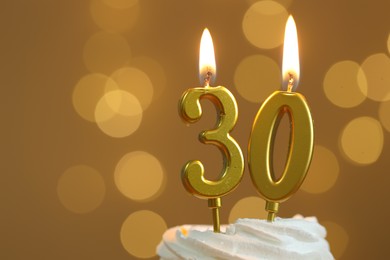 Photo of 30th birthday. Burning number shaped candles on cupcake against golden background with blurred lights, closeup and space for text. Bokeh effect