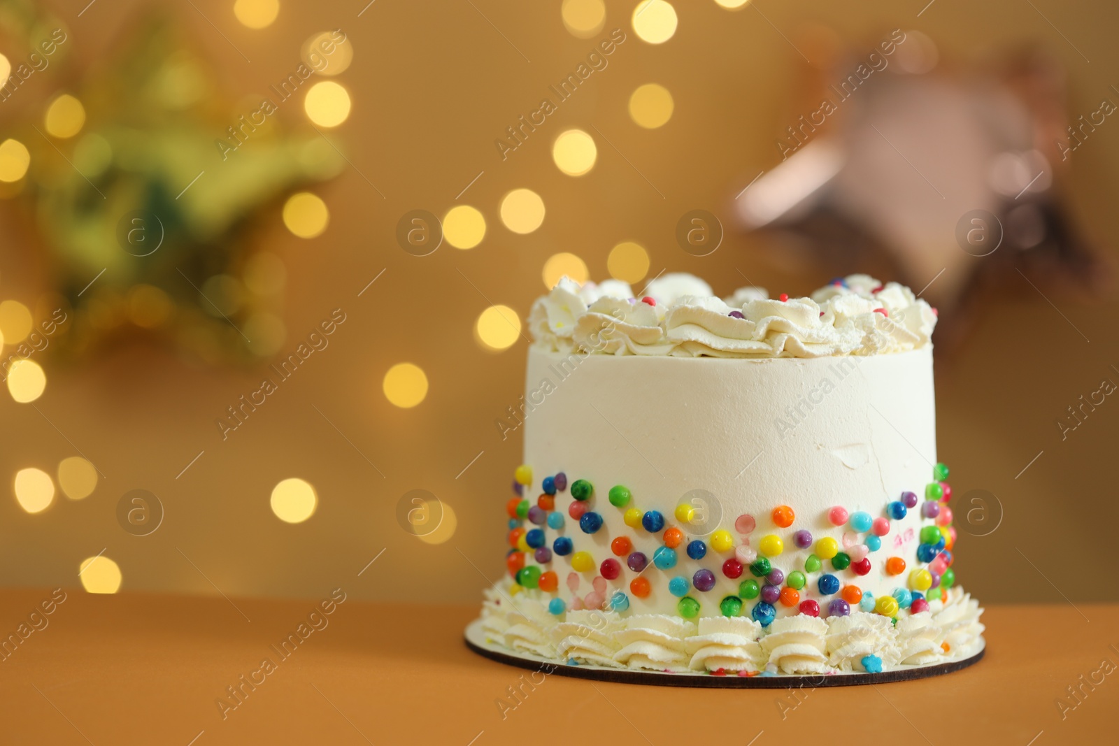 Photo of Tasty cake with cream and sprinkles on table against golden background with blurred lights, closeup. Space for text