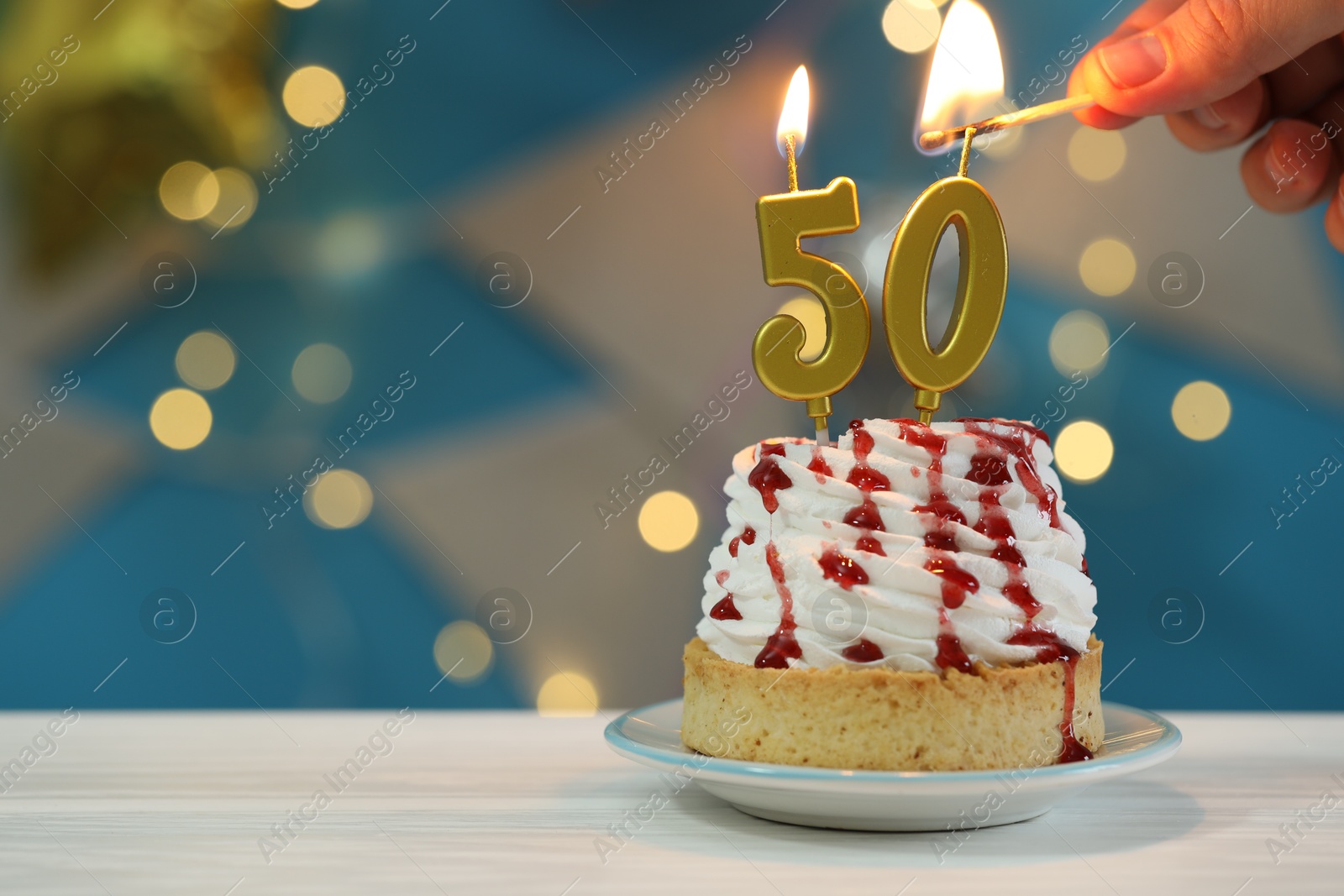 Photo of 50th birthday. Woman lighting number shaped candles on cupcake at white wooden table against blurred lights, closeup and space for text. Bokeh effect