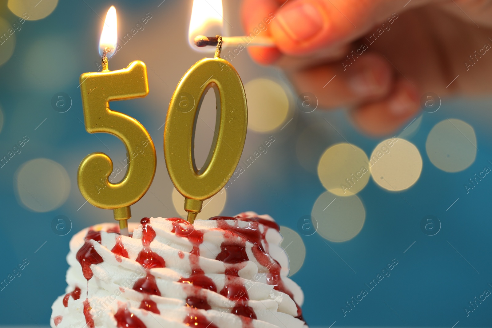 Photo of 50th birthday. Woman lighting number shaped candles on cupcake against blurred lights, closeup. Bokeh effect