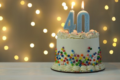 Photo of 40th birthday. Cake with burning number shaped candles on white wooden table against blurred background with lights, closeup and space for text. Bokeh effect