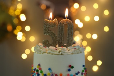 Photo of 50th birthday. Burning number shaped candles on cake against blurred background with lights, closeup. Bokeh effect