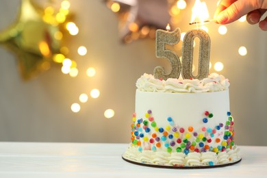 Photo of 50th birthday. Woman lighting number shaped candles on cake at white wooden table against blurred lights, closeup and space for text. Bokeh effect