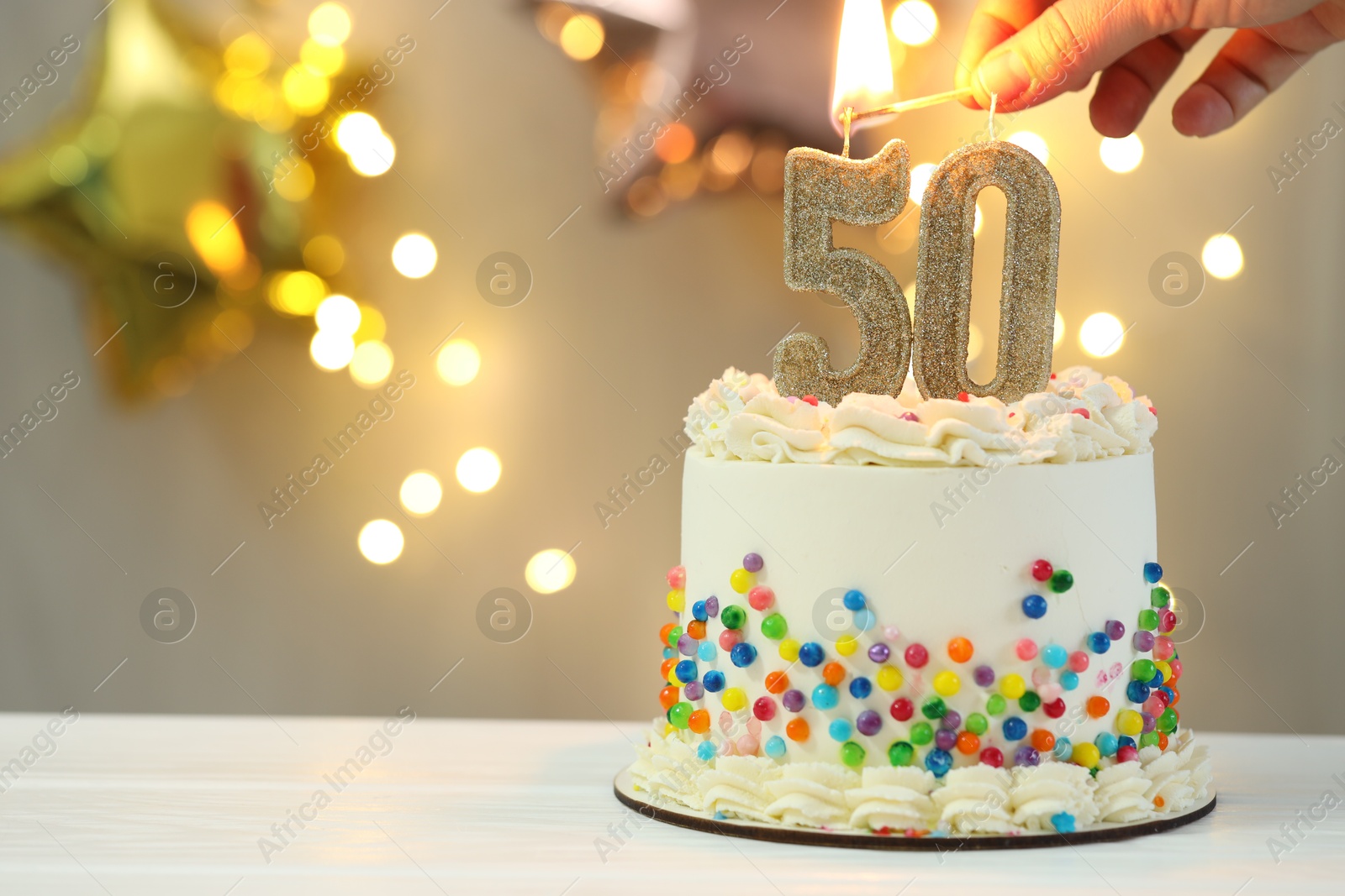 Photo of 50th birthday. Woman lighting number shaped candles on cake at white wooden table against blurred lights, closeup and space for text. Bokeh effect