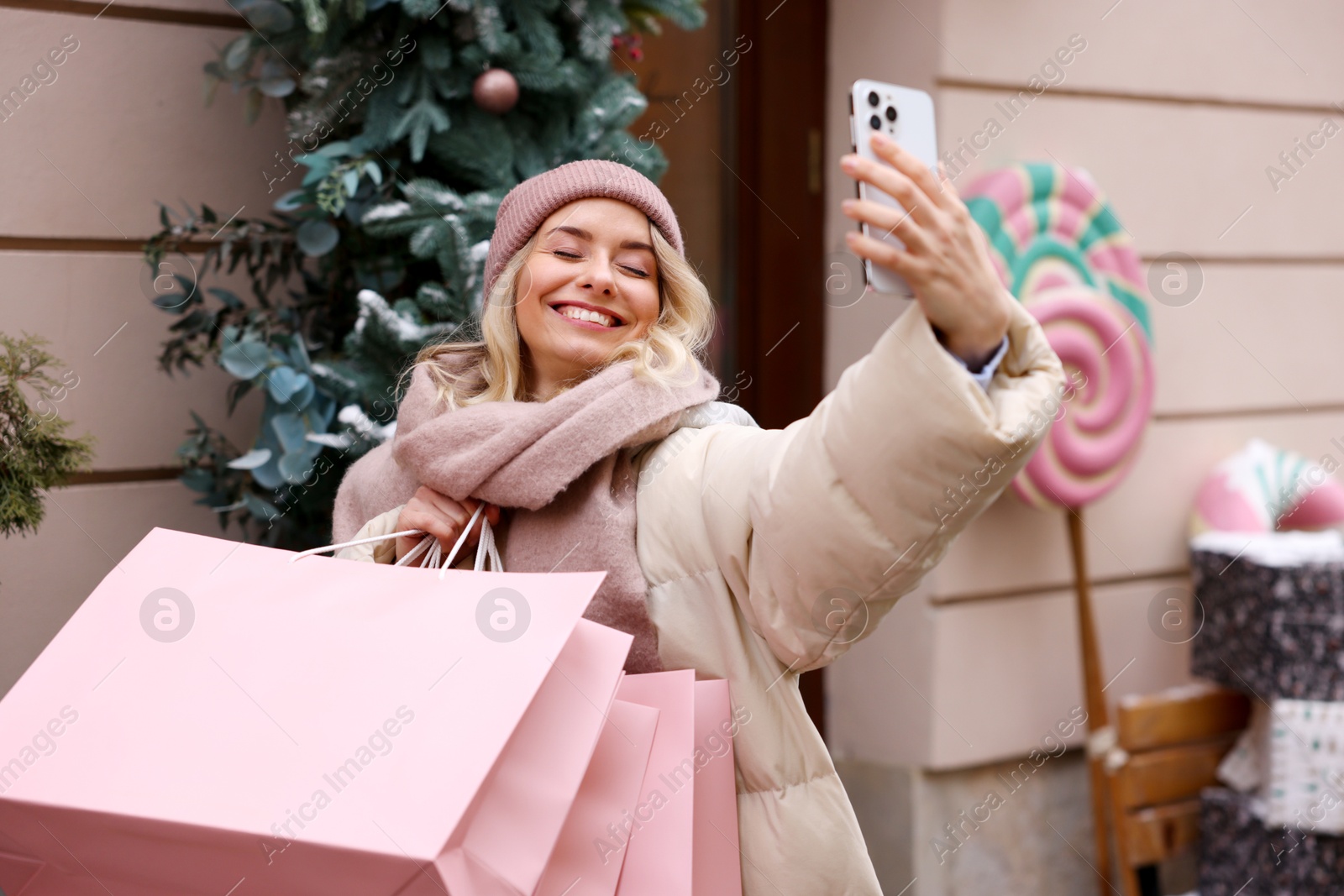 Photo of Happy woman with paper bags taking selfie indoors. Christmas season