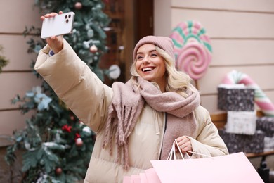 Photo of Happy woman with paper bags taking selfie indoors. Christmas season