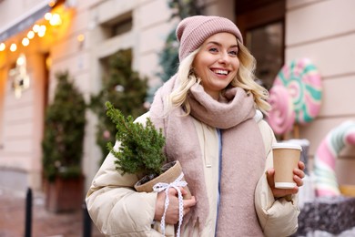 Photo of Happy woman with thuja tree and paper cup of coffee on city street, space for text. Christmas season