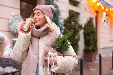 Photo of Happy woman with thuja tree and paper cup of coffee on city street, space for text. Christmas season