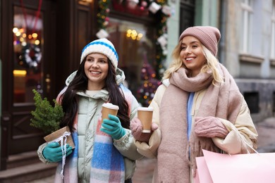 Photo of Happy friends with paper cups of coffee and thuja tree on city street. Christmas season