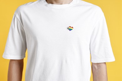 Photo of Man with heart shaped pin in colors of LGBT flag on yellow background, closeup