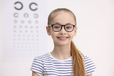 Photo of Little girl wearing glasses near vision test chart at ophthalmologist office, space for text