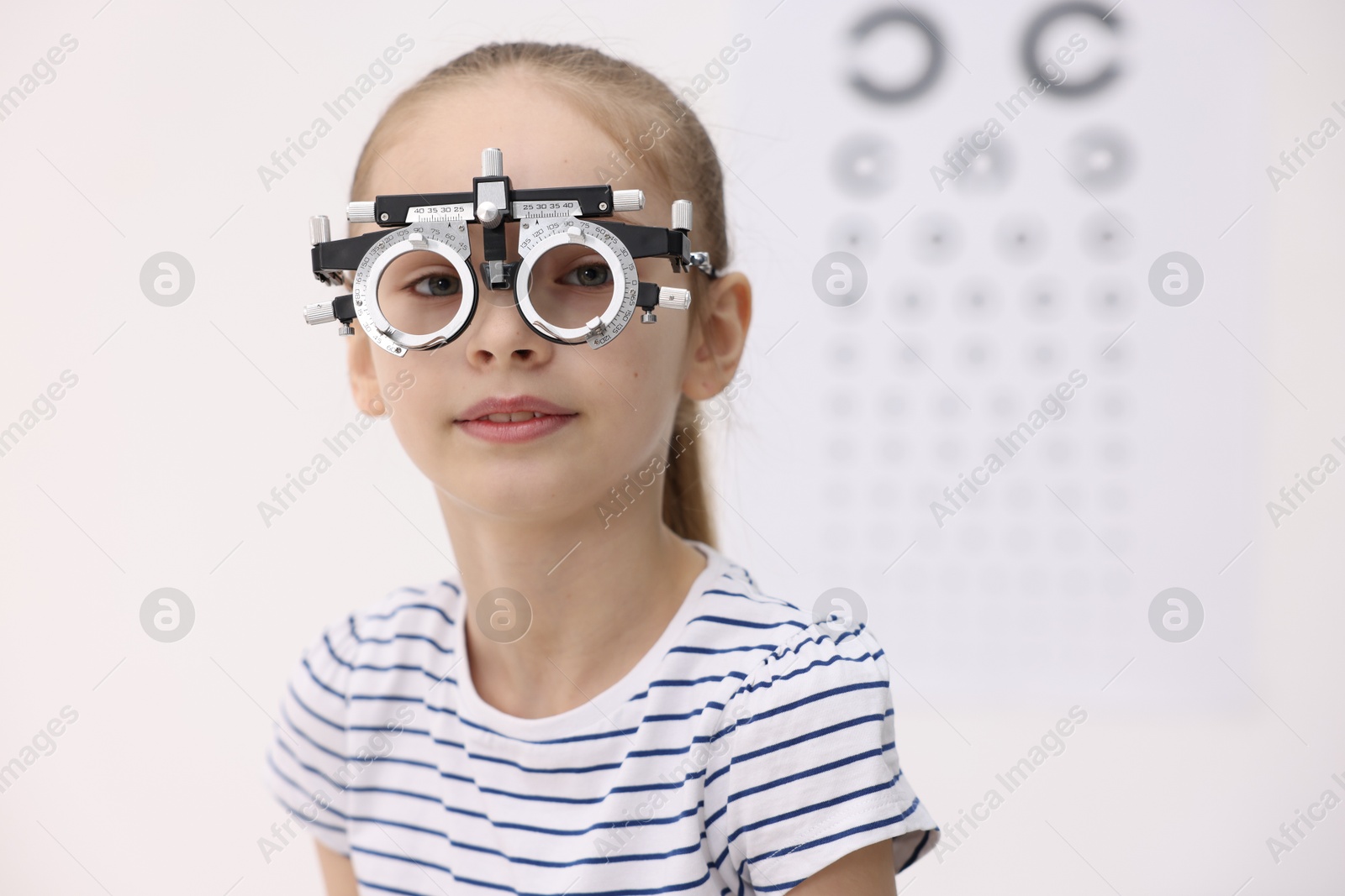 Photo of Little girl with trial frame near vision test chart at ophthalmologist office, space for text