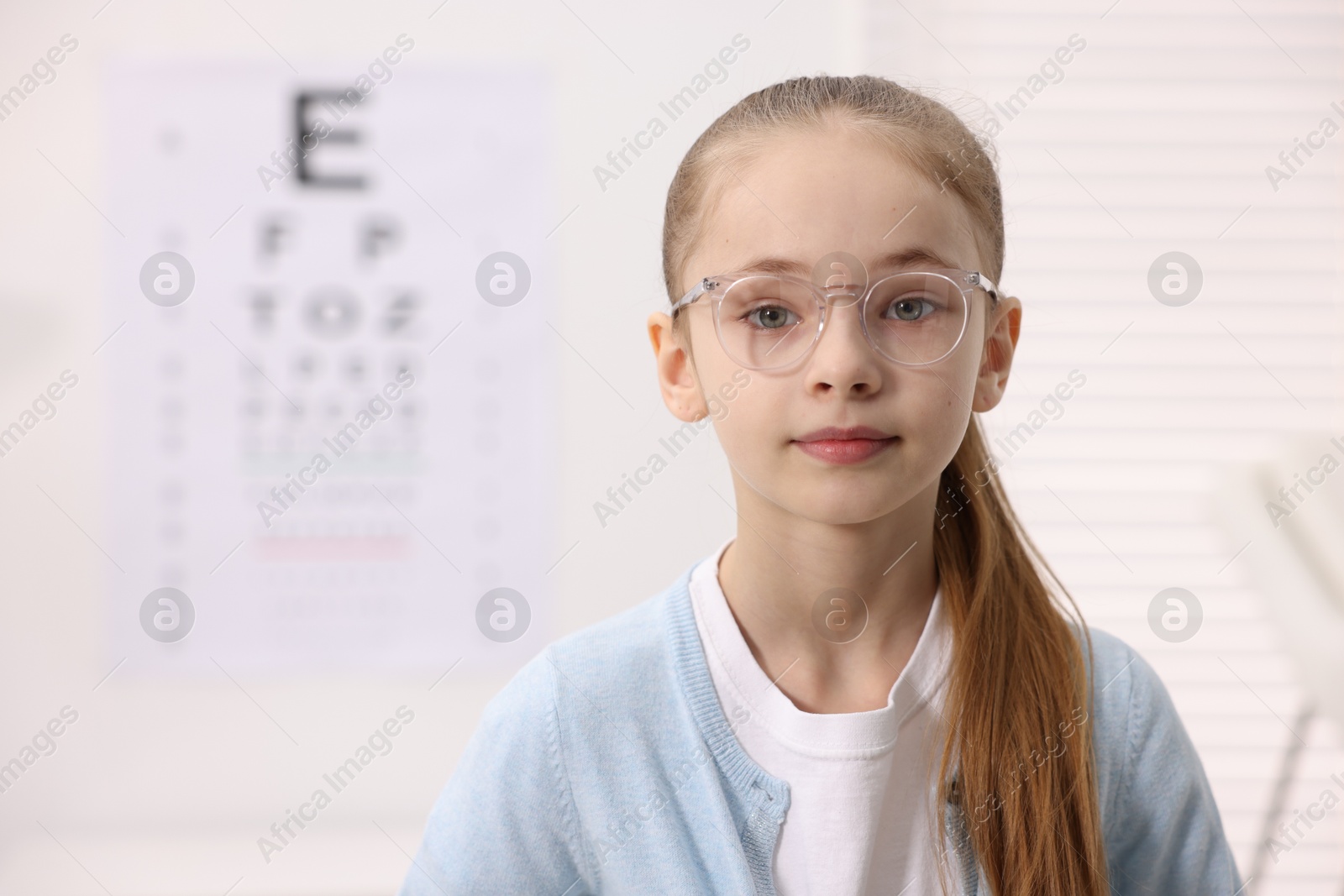 Photo of Little girl wearing glasses near vision test chart at ophthalmologist office, space for text