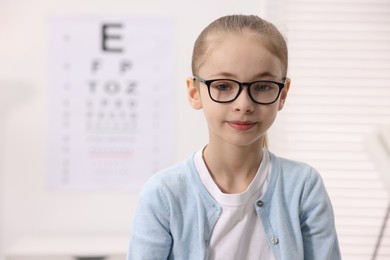 Photo of Little girl wearing glasses near vision test chart at ophthalmologist office, space for text