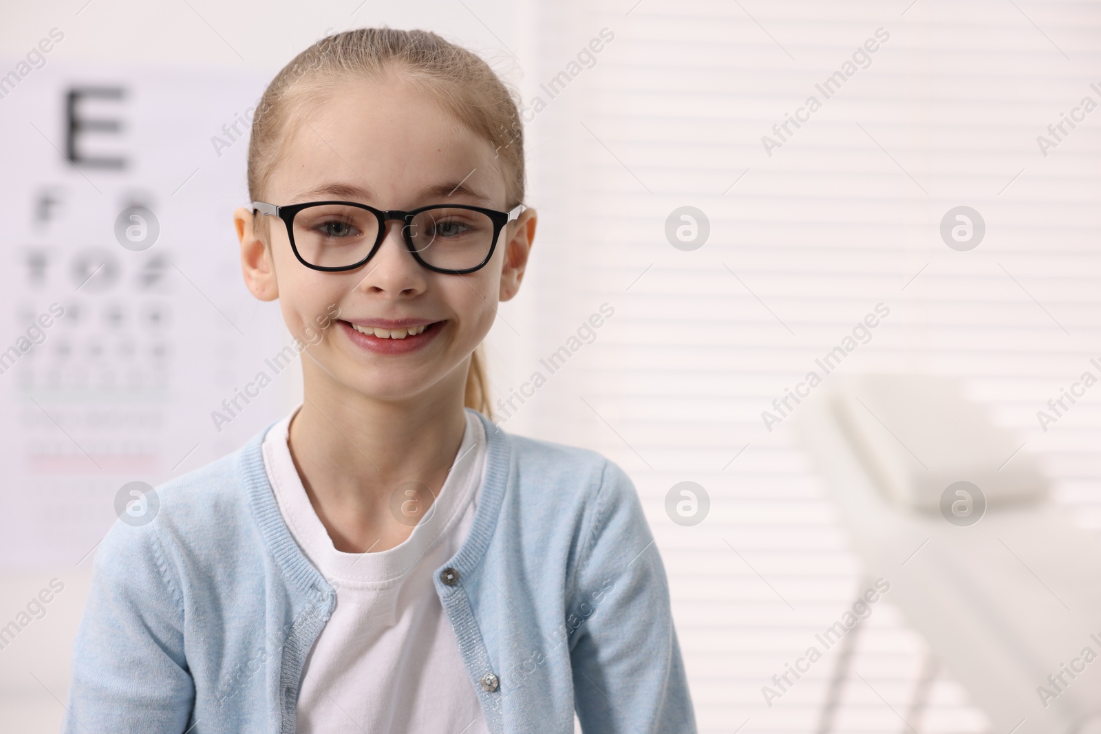 Photo of Little girl wearing glasses near vision test chart at ophthalmologist office, space for text