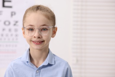 Photo of Little girl wearing glasses near vision test chart at ophthalmologist office, space for text