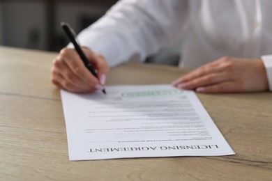 Photo of Woman signing licensing agreement document at wooden table indoors, closeup