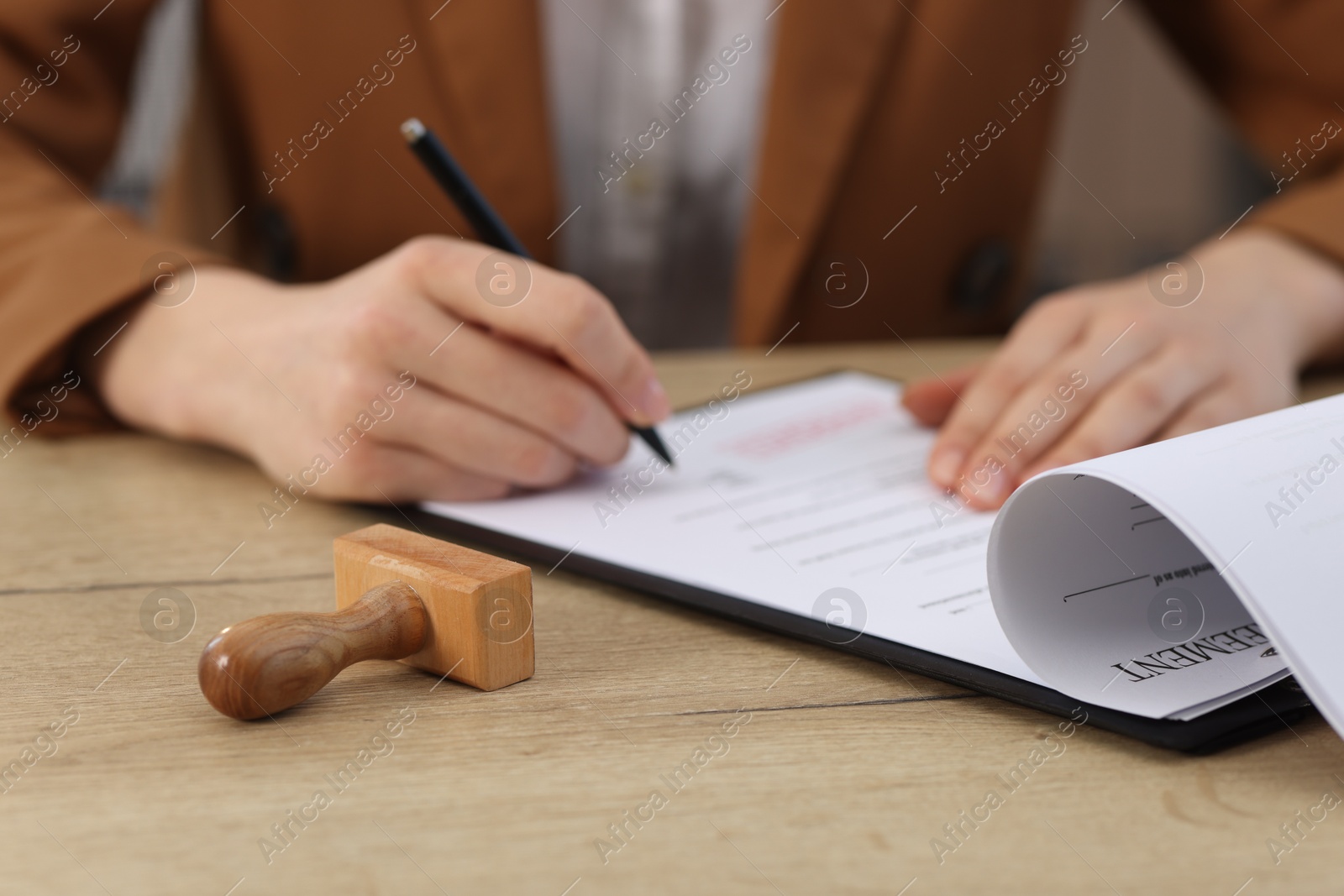 Photo of Woman signing licensing agreement document at wooden table indoors, focus on stamp