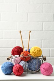 Photo of Balls of colorful yarn and knitting needles in metal basket on white wooden table, space for text