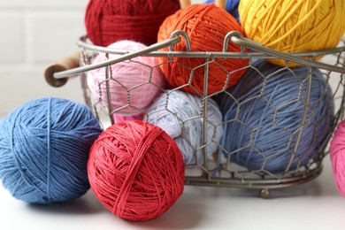 Photo of Balls of colorful yarn in metal basket on white wooden table, closeup