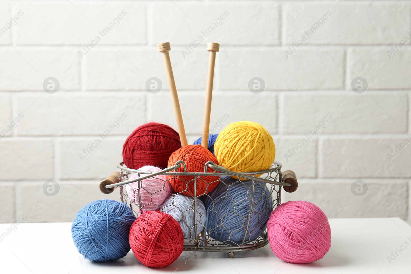 Balls of colorful yarn and knitting needles in metal basket on white wooden table Photo of Balls of colorful yarn and knitting needles in metal basket on white wooden table