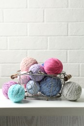 Balls of colorful yarn in metal basket on white wooden table, space for text