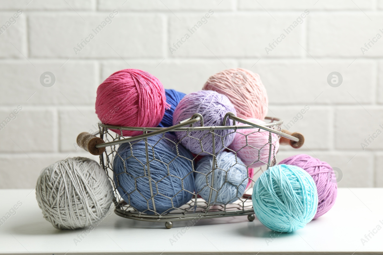 Photo of Balls of colorful yarn in metal basket on white wooden table, closeup