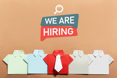 Image of We Are Hiring announcement. Paper shirts on pale brown background, top view. Human resource