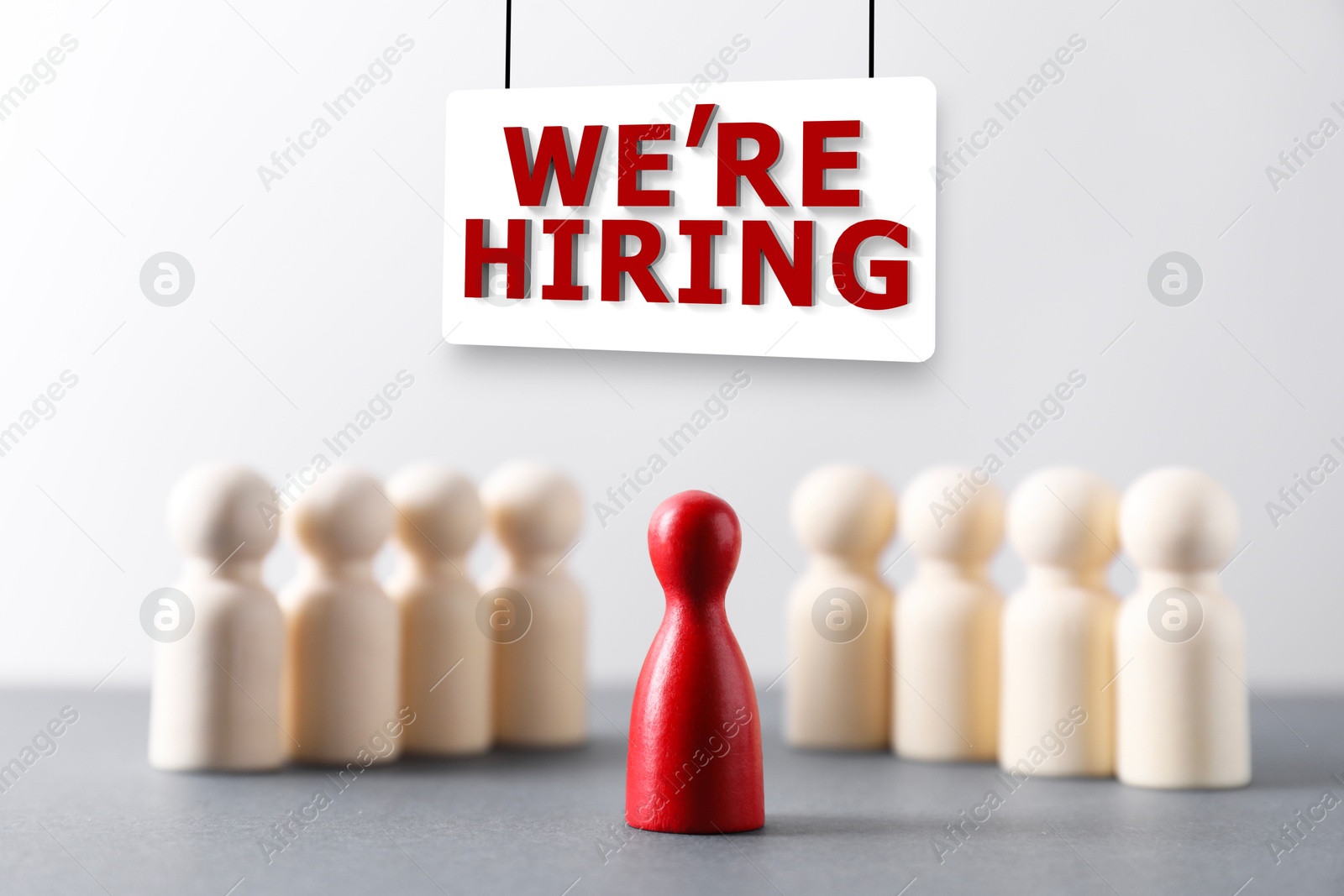 Image of We're Hiring sign board. Red figure among wooden ones on table. Human resource