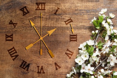 Image of Clock and blossoming branch with white flowers on wooden background, top view. Arrow showing time change one hour ahead in spring. Daylight saving time