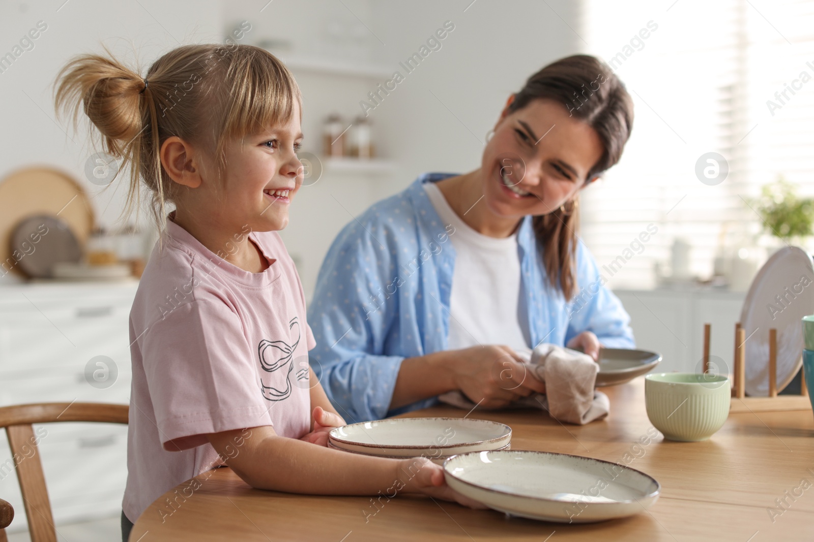 Photo of Little girl helping her mom wiping plates at table in kitchen