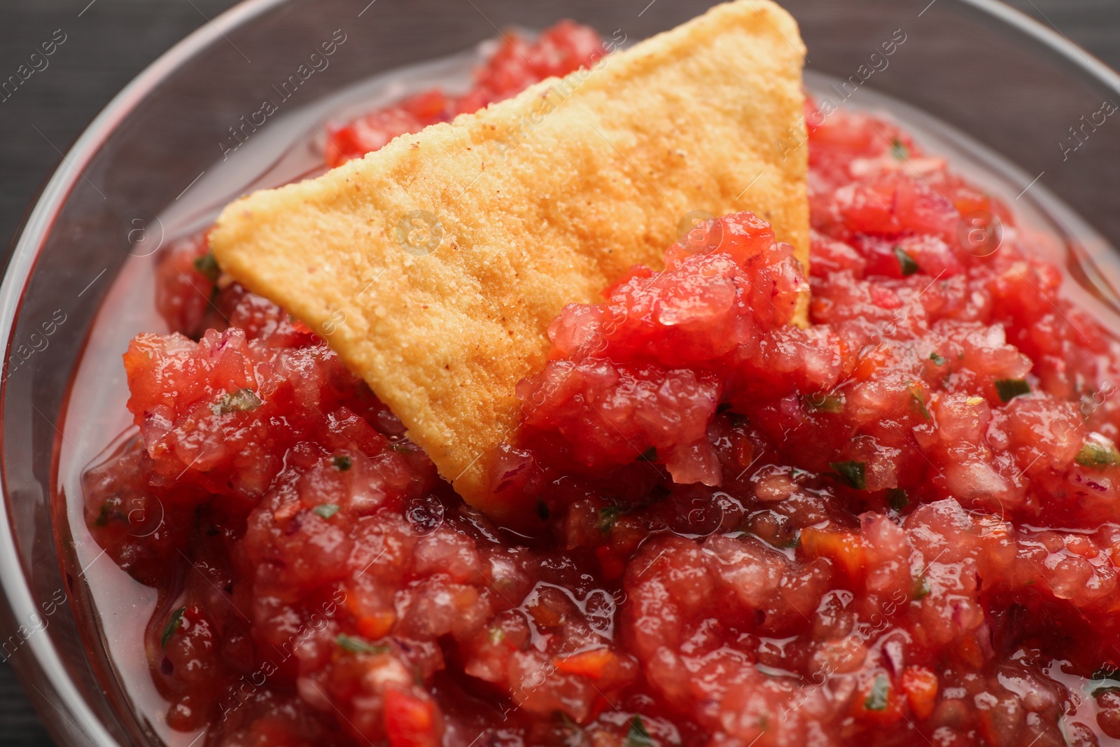 Photo of Spicy salsa sauce in glass bowl and nacho chip on table, closeup