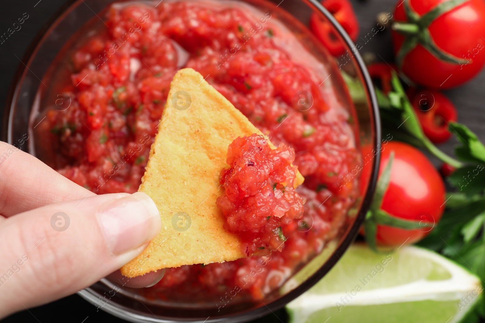 Photo of Woman dipping nacho chip into spicy salsa sauce at table, top view