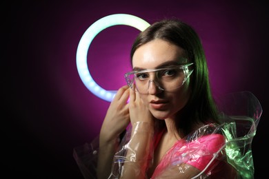 Photo of Stylish woman with ring lamp wearing transparent coat and glasses on dark purple background, space for text