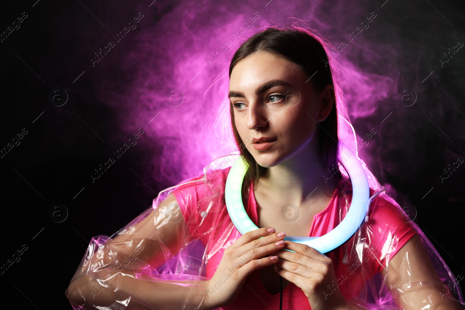 Photo of Beautiful woman with ring lamp wearing transparent coat on dark purple background in smoke