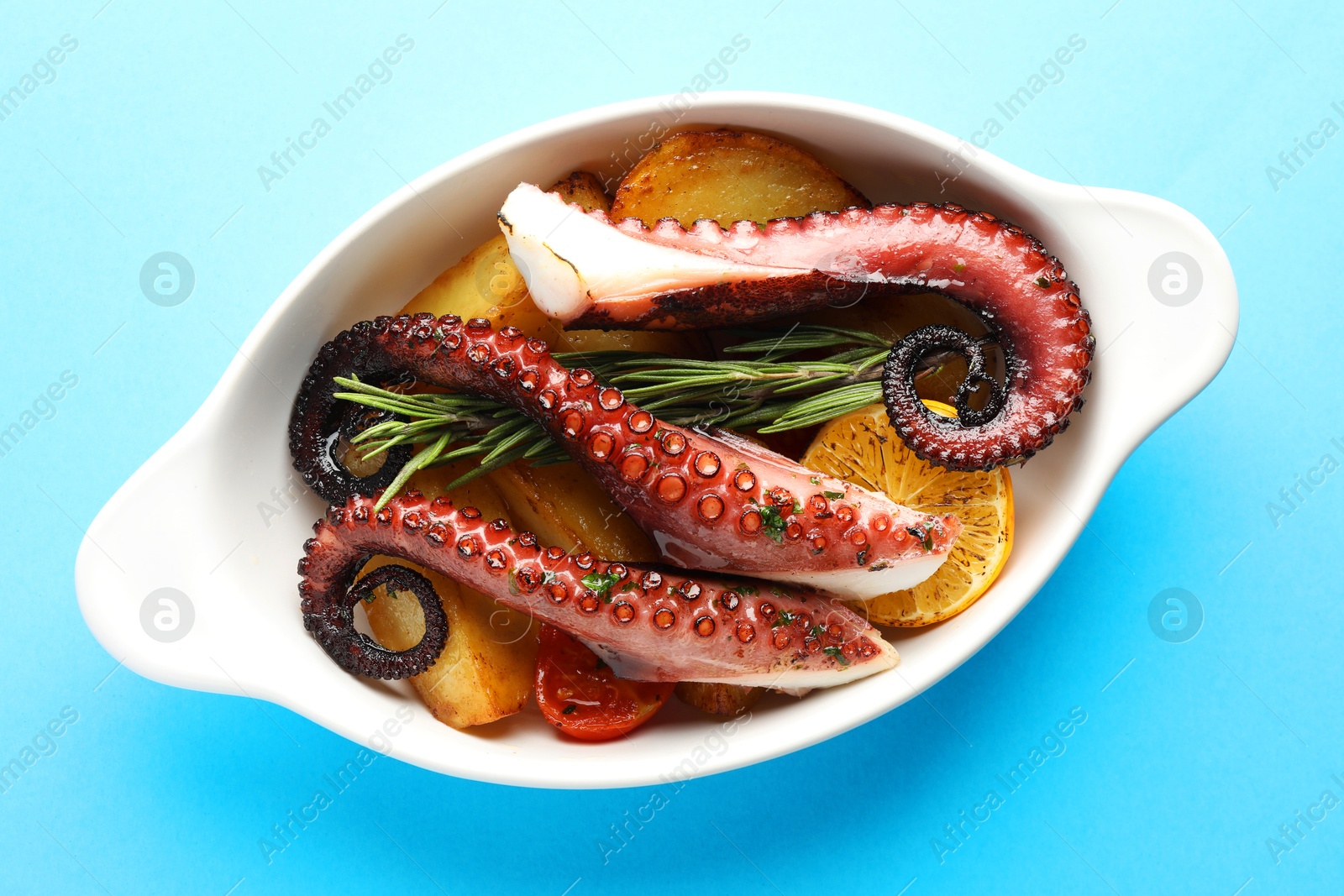 Photo of Fried octopus with vegetables, lemon and rosemary in baking dish on light blue background, top view