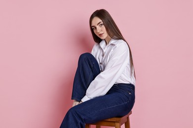 Beautiful young woman in stylish jeans sitting on stool against pink background