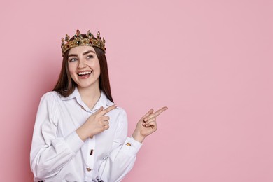 Photo of Happy woman in elegant crown pointing at something on pink background. Space for text