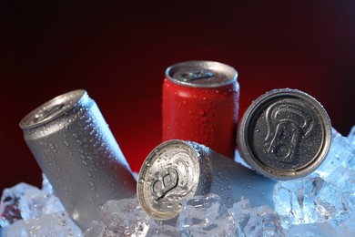 Photo of Refreshing drinks in cans on ice cubes against dark red background, closeup